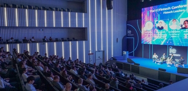 A large audience attentively watches a panel discussion at a fintech conference, with speakers displayed on a large screen above the stage.