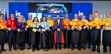 Group of academics in colorful robes standing on a stage at a formal event with a banner reading 'La fractura del orden internacional'.