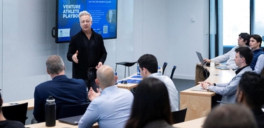 A speaker presenting in front of an audience in a modern classroom setting.