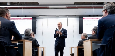 A man in a suit stands speaking in front of a group of seated individuals in a modern conference room.