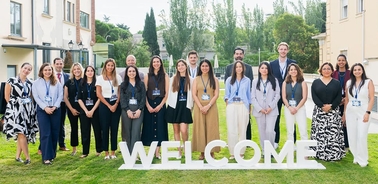 A group of diverse individuals stands together outdoors in front of a 'WELCOME' sign.