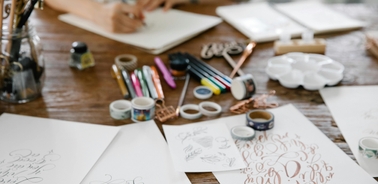 A person is drawing on a blank paper surrounded by various art supplies on a wooden table.
