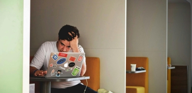 A man looks frustrated while using a laptop covered in stickers at a small table in a modern room.