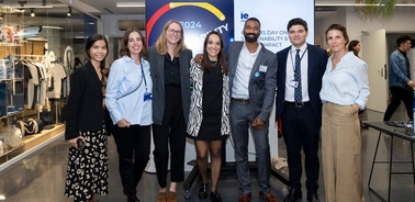 A group of seven people posing for a photo at a corporate event with a banner reading '2024 IE Day Sustainability and Impact' in the background.