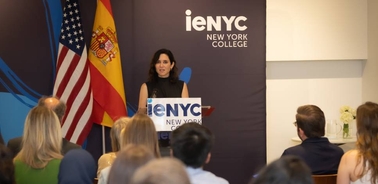 A woman speaks at a podium in front of flags representing the United States and Spain.