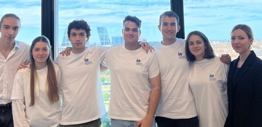 A group of seven young adults in white t-shirts labeled 'ie' standing indoors with a cityscape visible through a large window behind them.