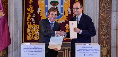 Two men in suits shaking hands while exchanging documents at a formal event with banners related to a university agreement.