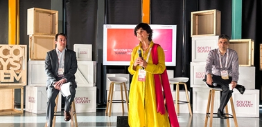 Three people participating in a panel discussion at a conference, with a woman in a yellow and red dress speaking and two men seated beside her.