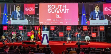 A business conference is taking place, with multiple speakers and large screens displaying a woman speaking.