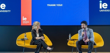 Two individuals are engaged in a discussion on stage at an IE University event, seated on yellow benches with a large 'THANK YOU' screen in the background.