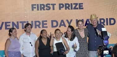 A group of seven people celebrating on stage at Venture Day Madrid, with a 'First Prize' banner in the background.