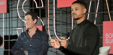 Two men sitting on a panel discussion at a conference, with one speaking and the other listening.