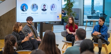 A panel discussion featuring four speakers on a stage with a large screen displaying their profiles.