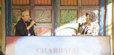 Two women engaged in a conversation on a stage with ornate background and a sign reading 'CHARBAGH'.