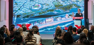 A speaker presenting to an audience with a large screen displaying a cityscape in the background.