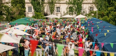 A vibrant outdoor market scene with numerous people browsing under colorful tents and strings of flags.