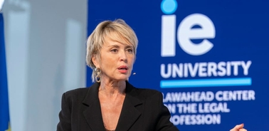 A woman speaks passionately at a podium with a banner of IE University in the background.