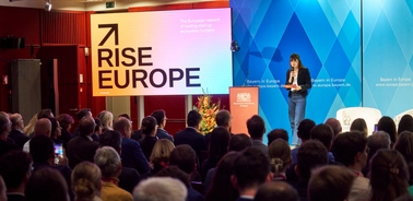 A woman is giving a presentation at a conference with a large audience in a room decorated with red and blue geometrical designs.