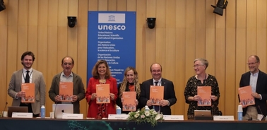 A group of seven people standing behind a desk at a UNESCO event, holding orange booklets and smiling.