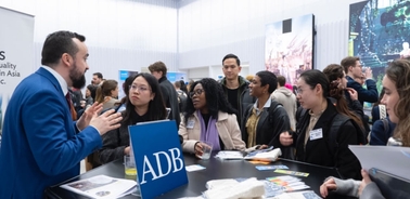 A man in a suit is speaking to a group of young adults at a career fair booth labeled 'ADB'.