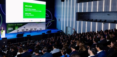 A speaker is presenting on stage at a conference with a large audience in attendance.