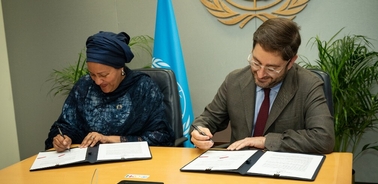Two individuals, one woman and one man, are signing documents at a desk with a UN logo visible in the background.