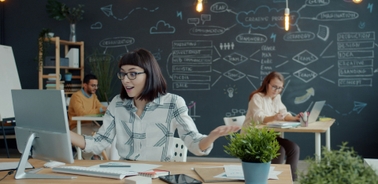 A woman at a desk reacts with excitement while working on a computer in a creative office environment.