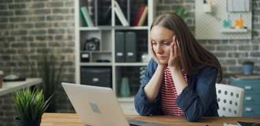 A young woman is sitting at a desk with her hands on her head, looking stressed while working on a laptop.