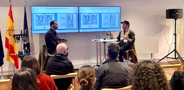 Two speakers are engaged in a discussion in front of an audience, with screens displaying visuals behind them.