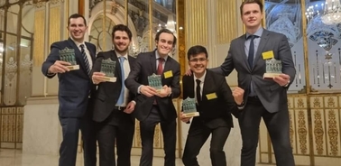 Five men in formal attire holding books and smiling in a grand hallway.