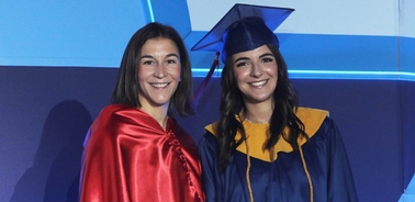 Two women in graduation attire, one in red and the other in blue, smiling at a graduation ceremony.