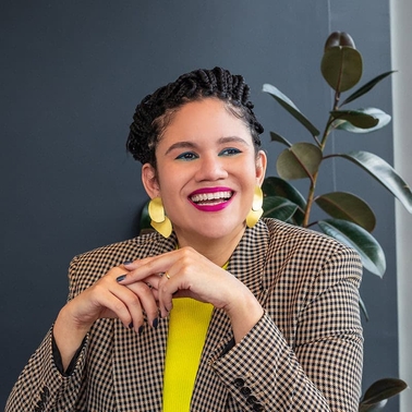 A smiling woman wearing a plaid blazer and bright yellow top poses against a backdrop of indoor plants.
