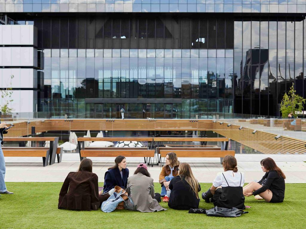 A group of young people are sitting on a lawn in front of a modern building talking and sharing.