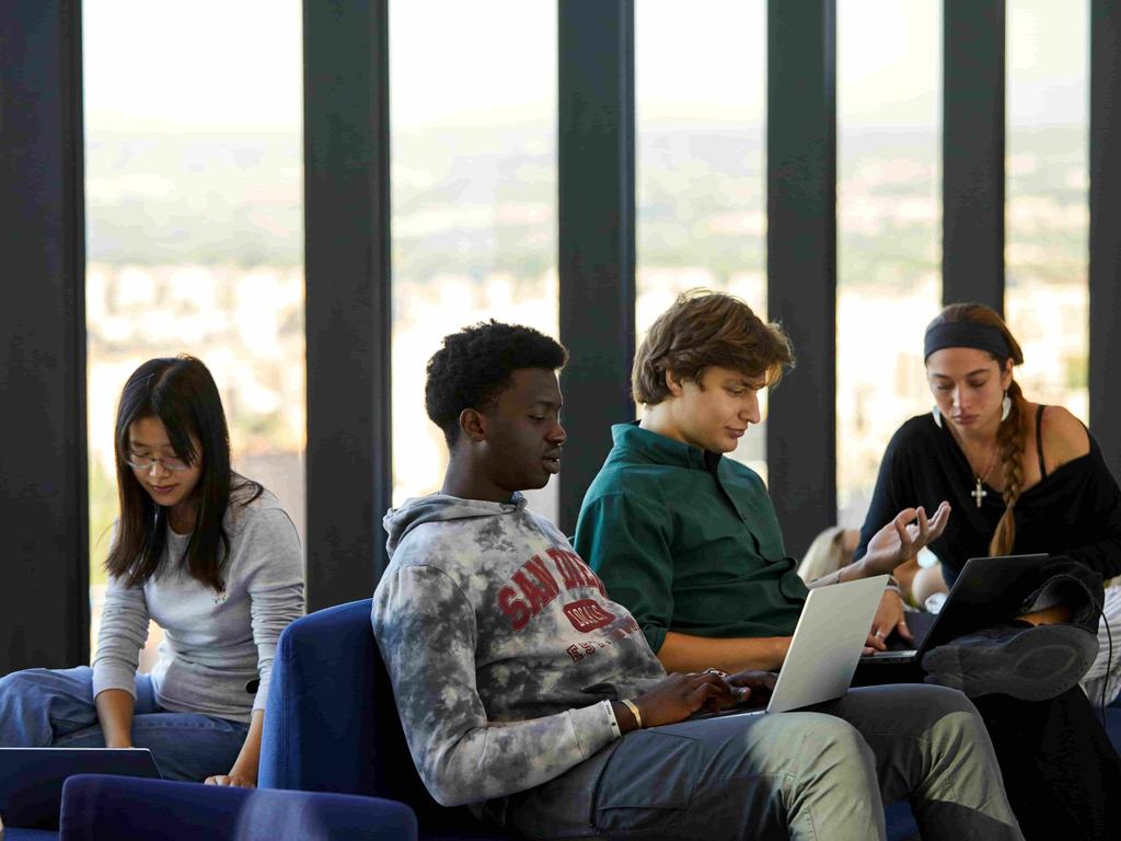 A group of young people working collaboratively on laptops in a modern space with large windows.