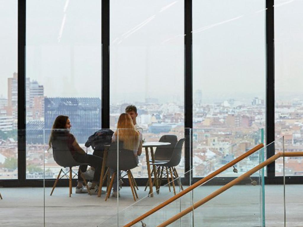 Three people are sitting at a table in a modern office with large windows overlooking the city.