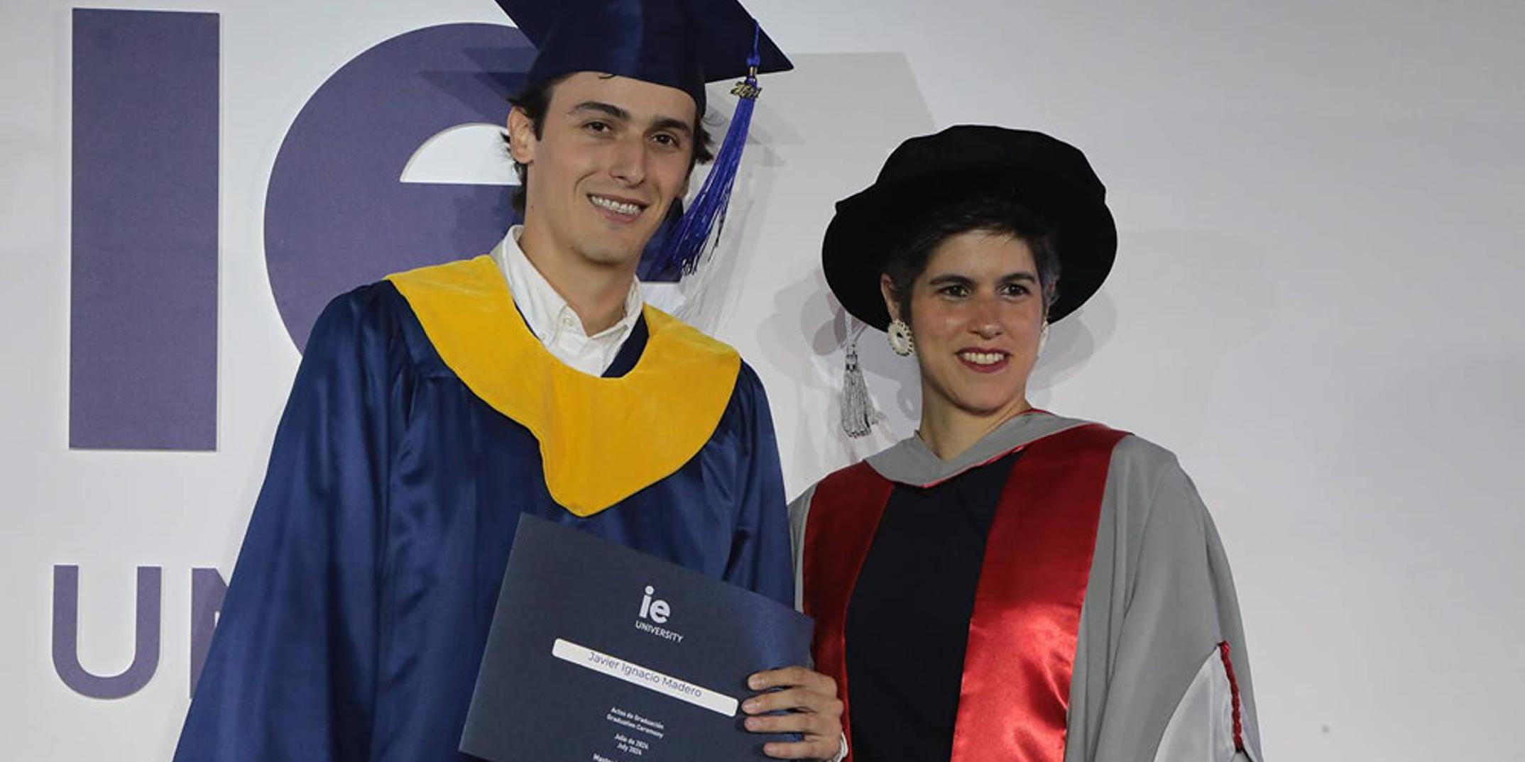 A young man in a graduation gown poses with a woman in academic regalia at a graduation ceremony.