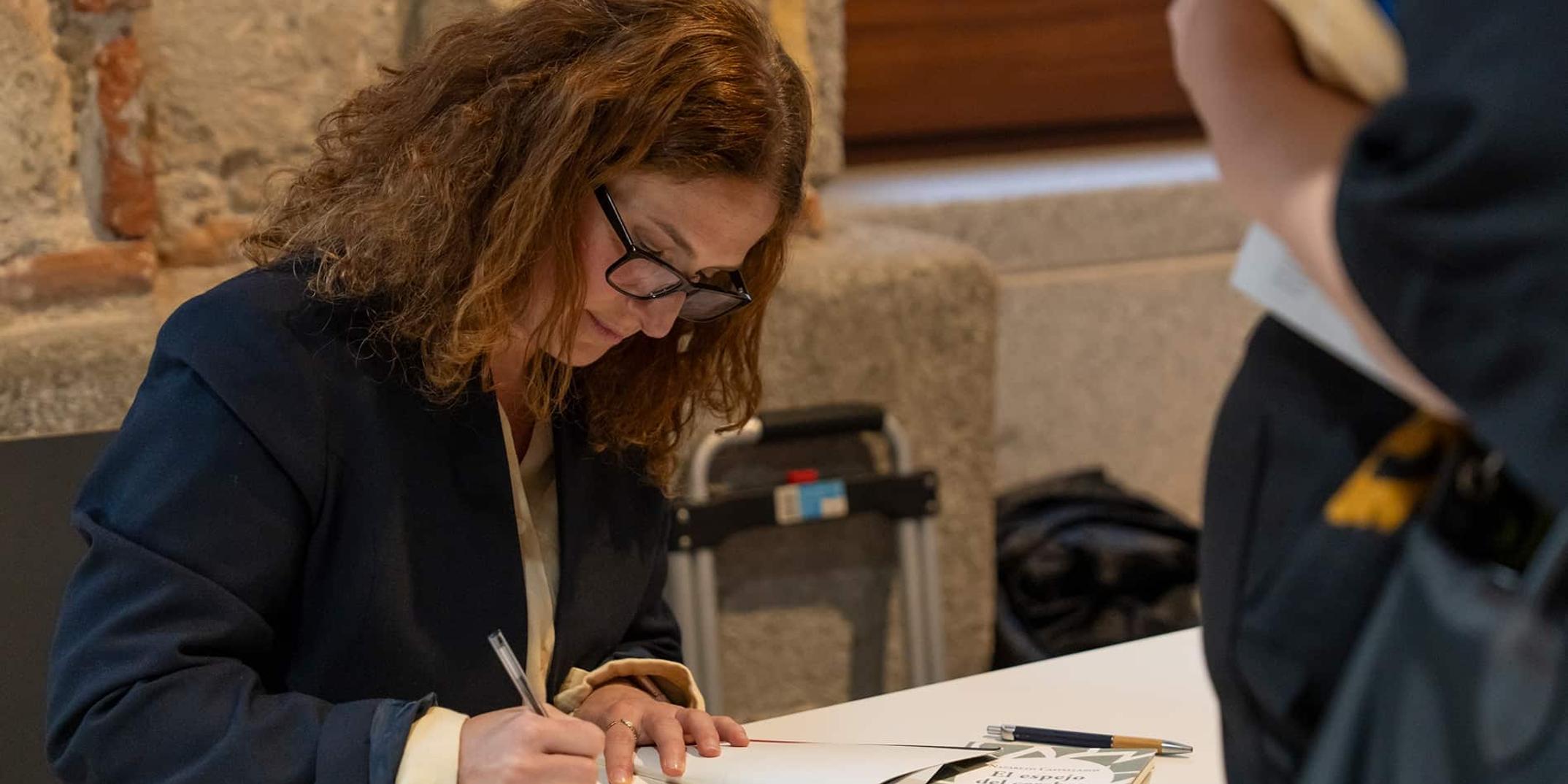 A woman is signing a book at a table during an event.