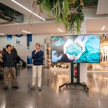 A group of people gathered around a presentation screen displaying hands holding green material in a modern indoor setting.