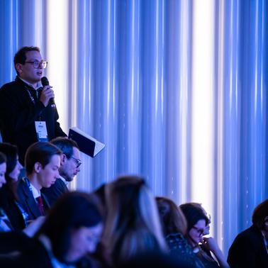 A person is speaking at a conference while others are seated in the audience.
