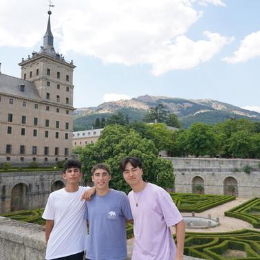 Three young men standing in front of a historic building with gardens and mountains in the background.