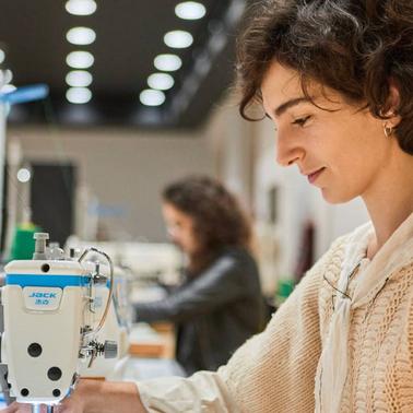 A woman operating a sewing machine in a workshop setting with other people in the background.
