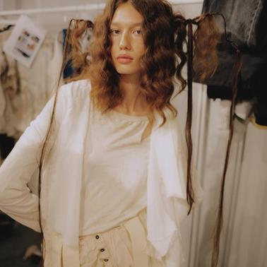 A model with curly hair styled into two long, wavy strands stands in a fashion setting.