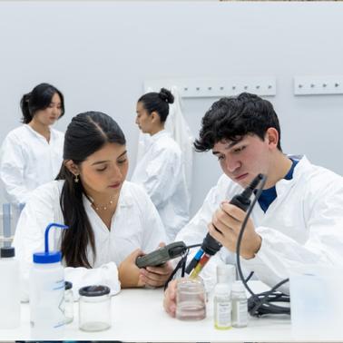 Two students are working together in a laboratory, one using a pipette and the other looking at a phone.