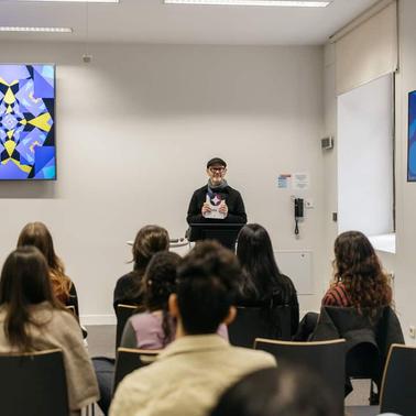 A speaker presents to an audience in a well-lit room decorated with colorful artwork.