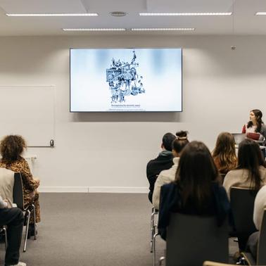 A presenter speaks to an audience in a modern conference room with a large screen displaying an image.