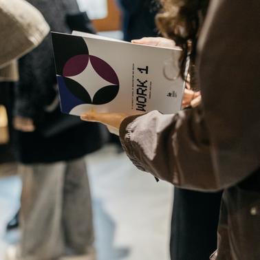 A person is holding a booklet titled 'WORK 1' in a crowded setting.