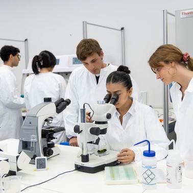 A group of students in white lab coats are engaged in a science experiment involving a microscope.