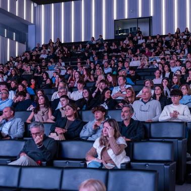 A large audience is seated in a modern auditorium, attentively watching a presentation.