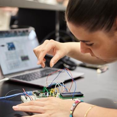 A person is focused on assembling a circuit with wires and components while working on a laptop.