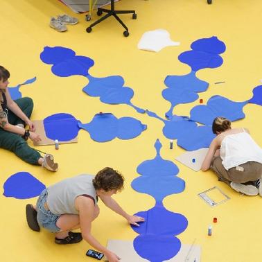 Three artists are painting large blue shapes on a yellow floor.